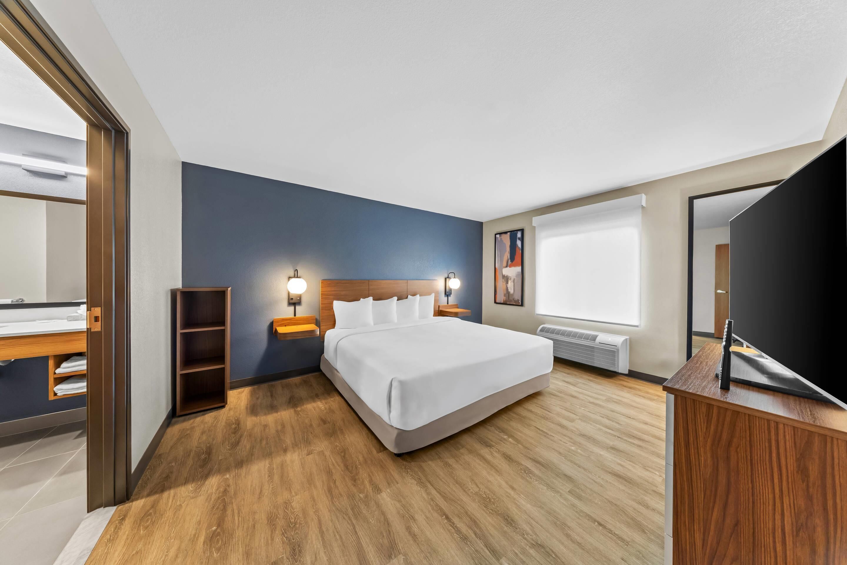 A modern hotel room with a large bed, wooden flooring, a built-in wardrobe, bedside lamps, and a large window.