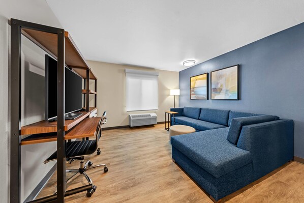 A modern hotel room with a wooden desk, a blue sofa, a standing desk with a TV, and a large window. - Everhome Suites Ontario Airport (Ontario)