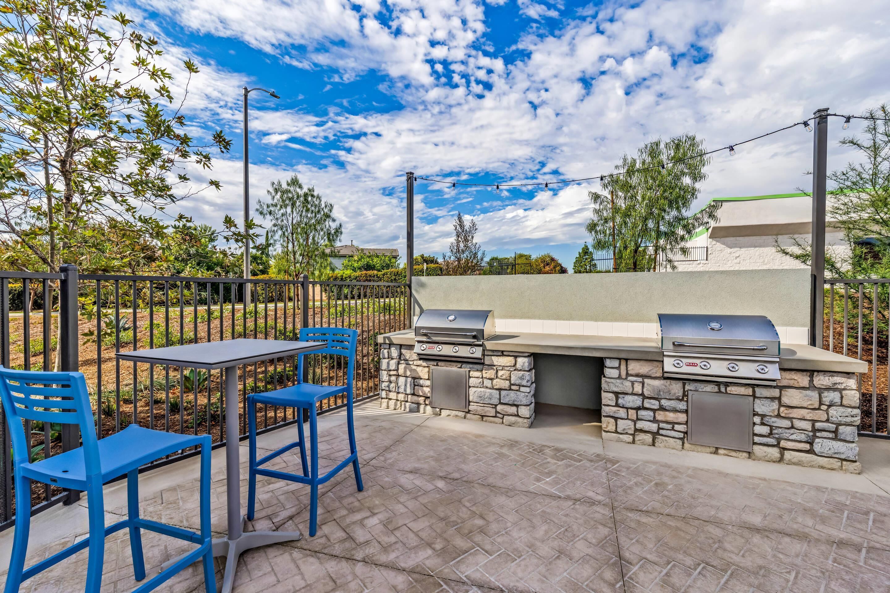 An outdoor patio with a stone barbecue grill, two blue chairs, and a table.