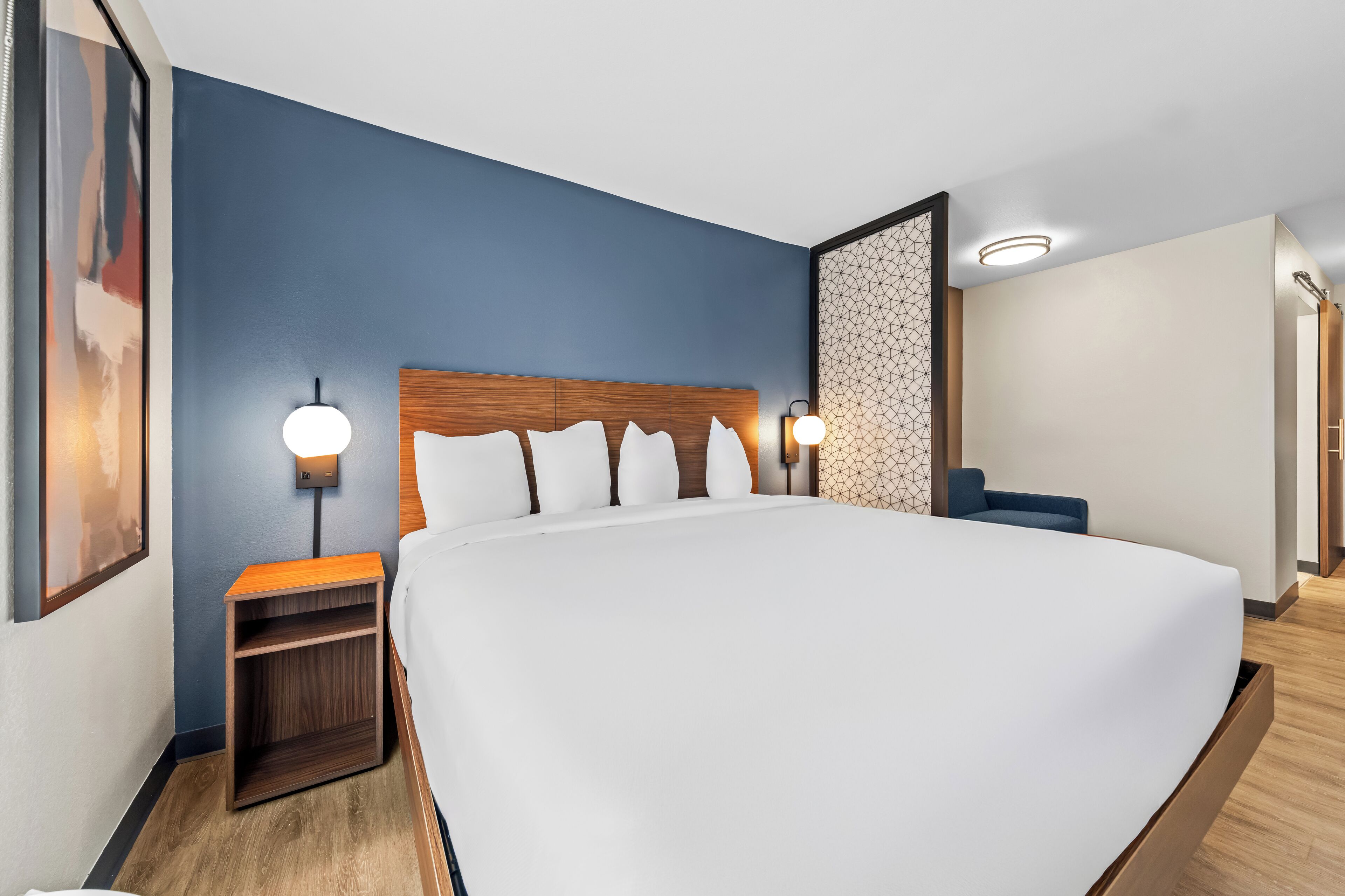 Een moderne hotelkamer met een groot bed, een houten hoofdbord, twee nachtkastjes met lampen en een scheidingswand met een geometrisch patroon.