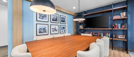 A conference room with a wooden table, grey upholstered chairs, a large flat-screen TV, and a bookshelf with books.