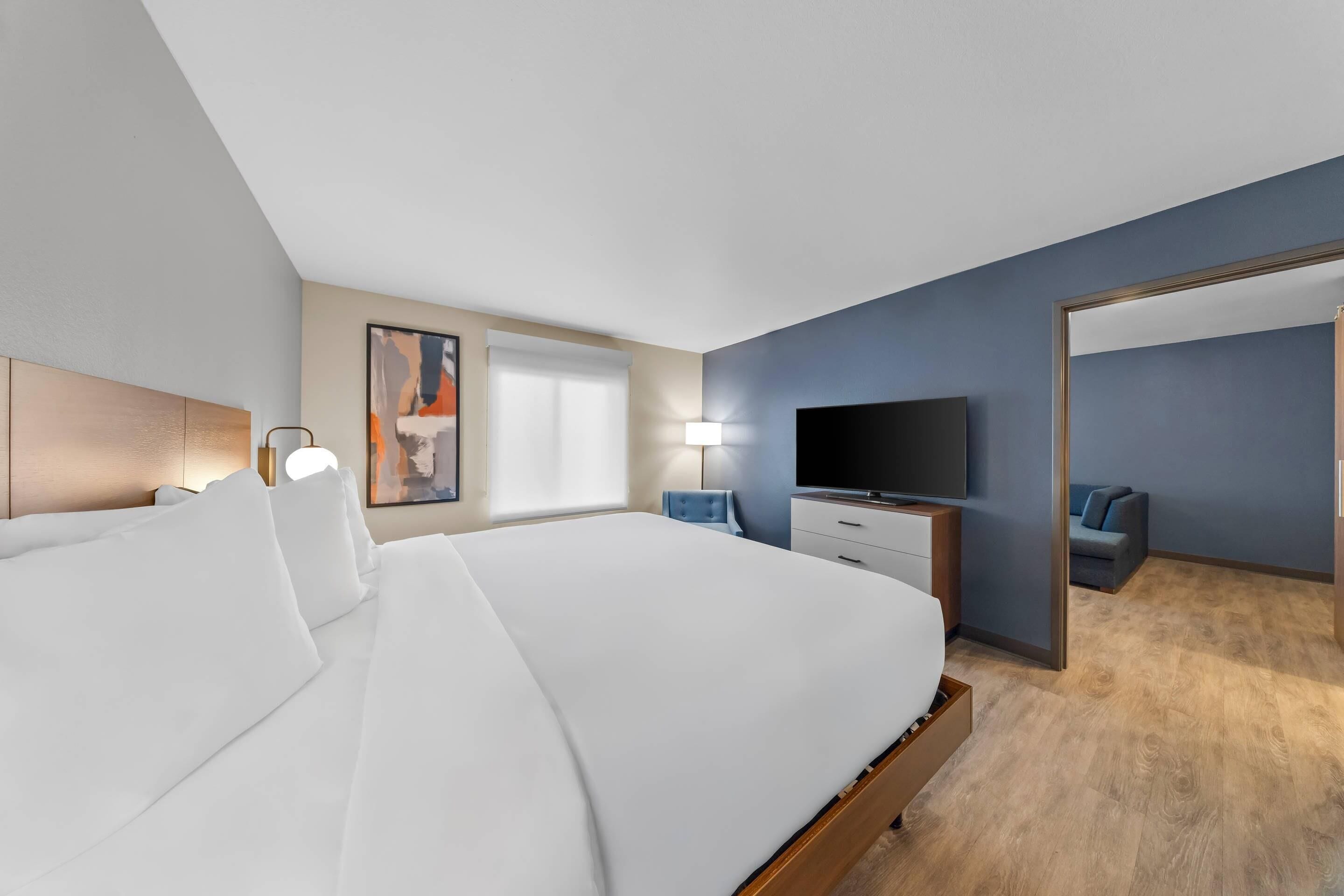A modern hotel room with a large bed, a flat-screen TV, a desk with a lamp, and a view of the hallway.