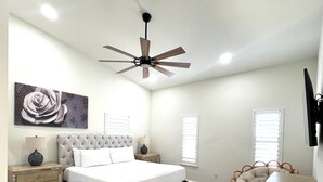 A bedroom with a tufted headboard bed, a ceiling fan, a large floral artwork, a nightstand with drawers, a bedside table, a wooden dresser, and a patterned rug.