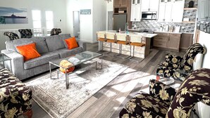 A spacious living room with a grey sofa, orange cushions, a glass coffee table, and floral-patterned armchairs. A kitchen with white cabinets and a star-shaped pendant light is visible in the background.
