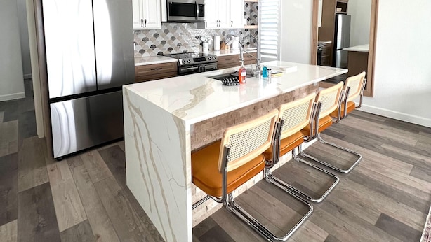 A modern kitchen with a central island, stainless steel appliances, and a wooden ceiling fan.