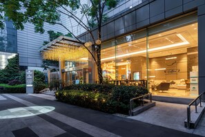 Un edificio moderno con fachada de vidrio, una cafetería visible en su interior y un área exterior bien cuidada con un árbol y arbustos.
