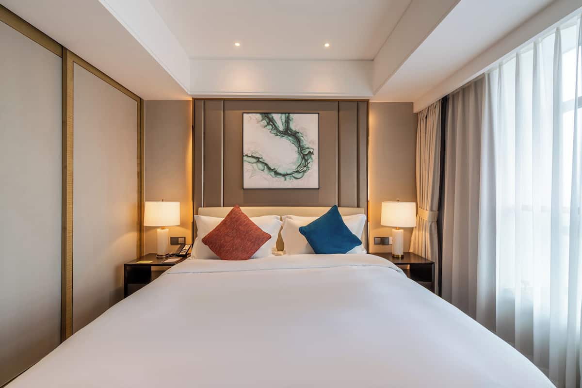 Habitación de hotel con una cama grande, dos mesitas de noche con lámparas, un cuadro en la pared y una ventana con cortinas.
