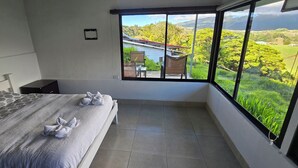 A bedroom with a large window, a bed with towels, a small table, and a chair.