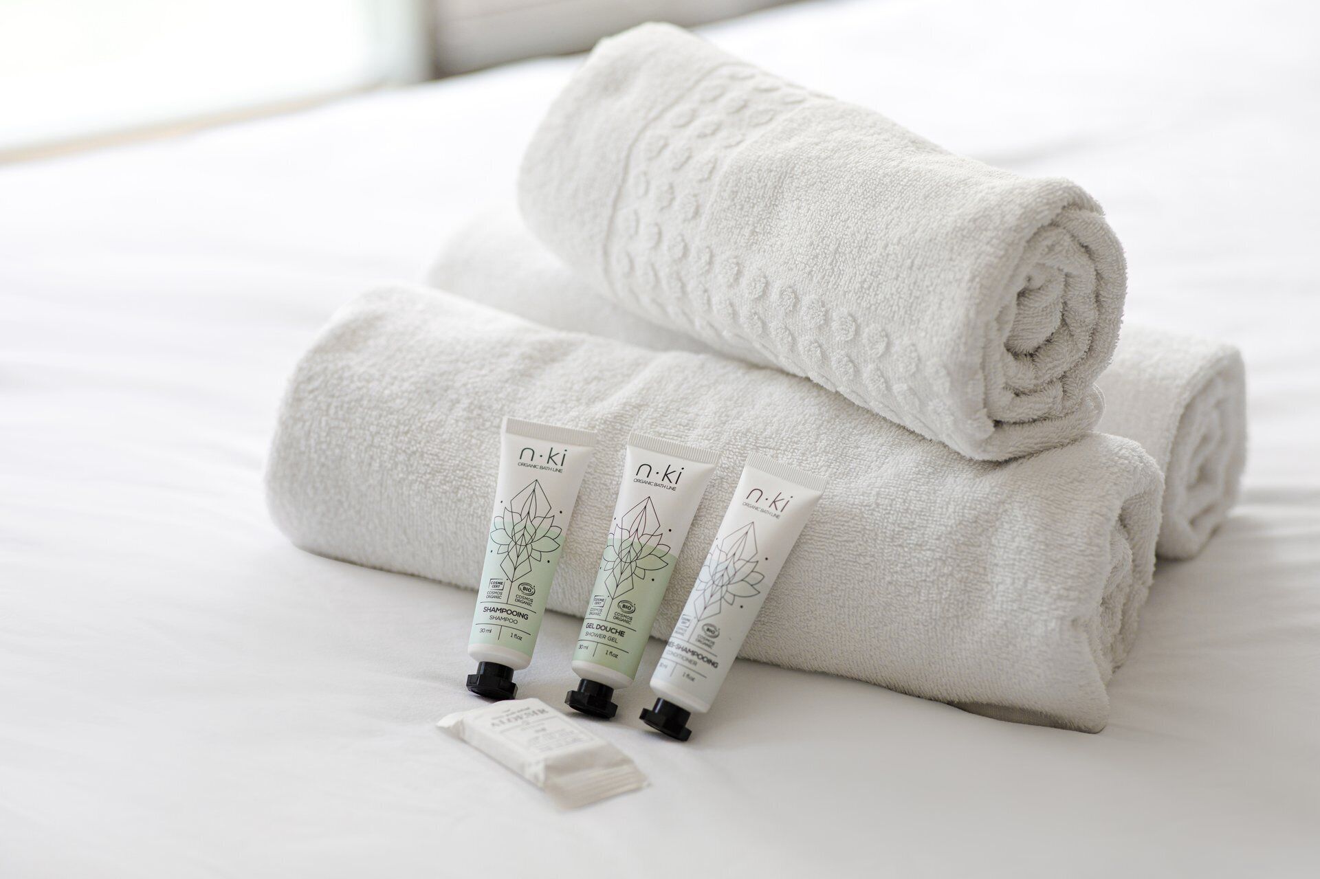 Ein ordentlich bezogenes Bett mit aufgerollten weißen Handtüchern und zwei Flaschen Shampoo und Duschgel aus der n.ki Organic Bath Line.