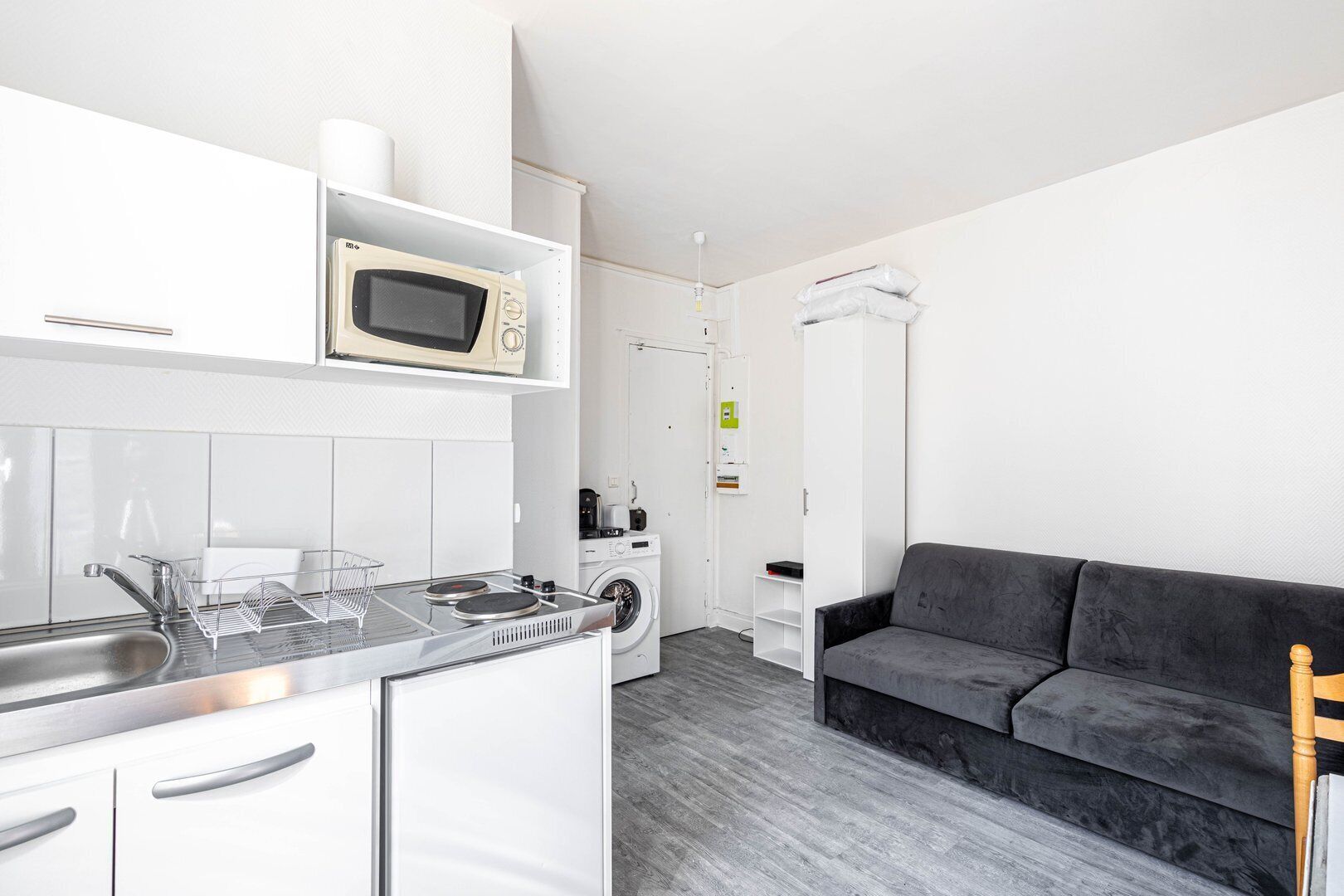 A compact kitchen with a microwave, stove, and washing machine, a gray sofa, and a wooden chair.