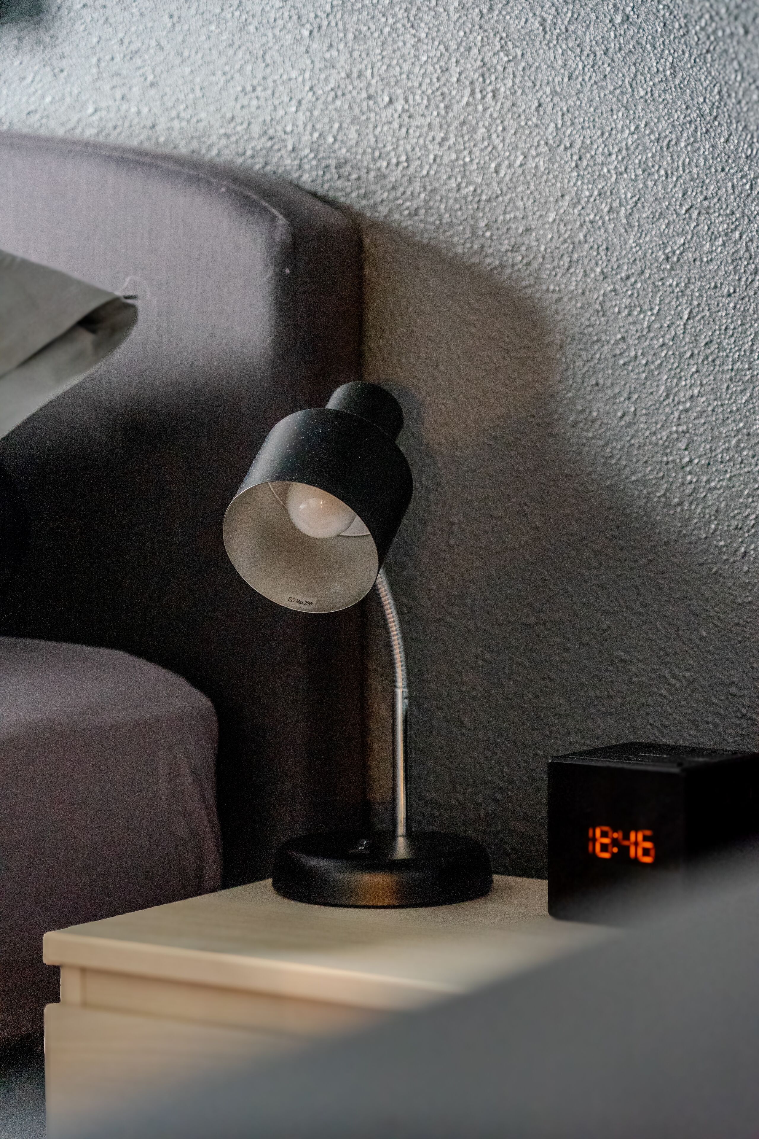 Uma mesa de cabeceira com uma luminária de mesa preta e um relógio digital mostrando 18:46.