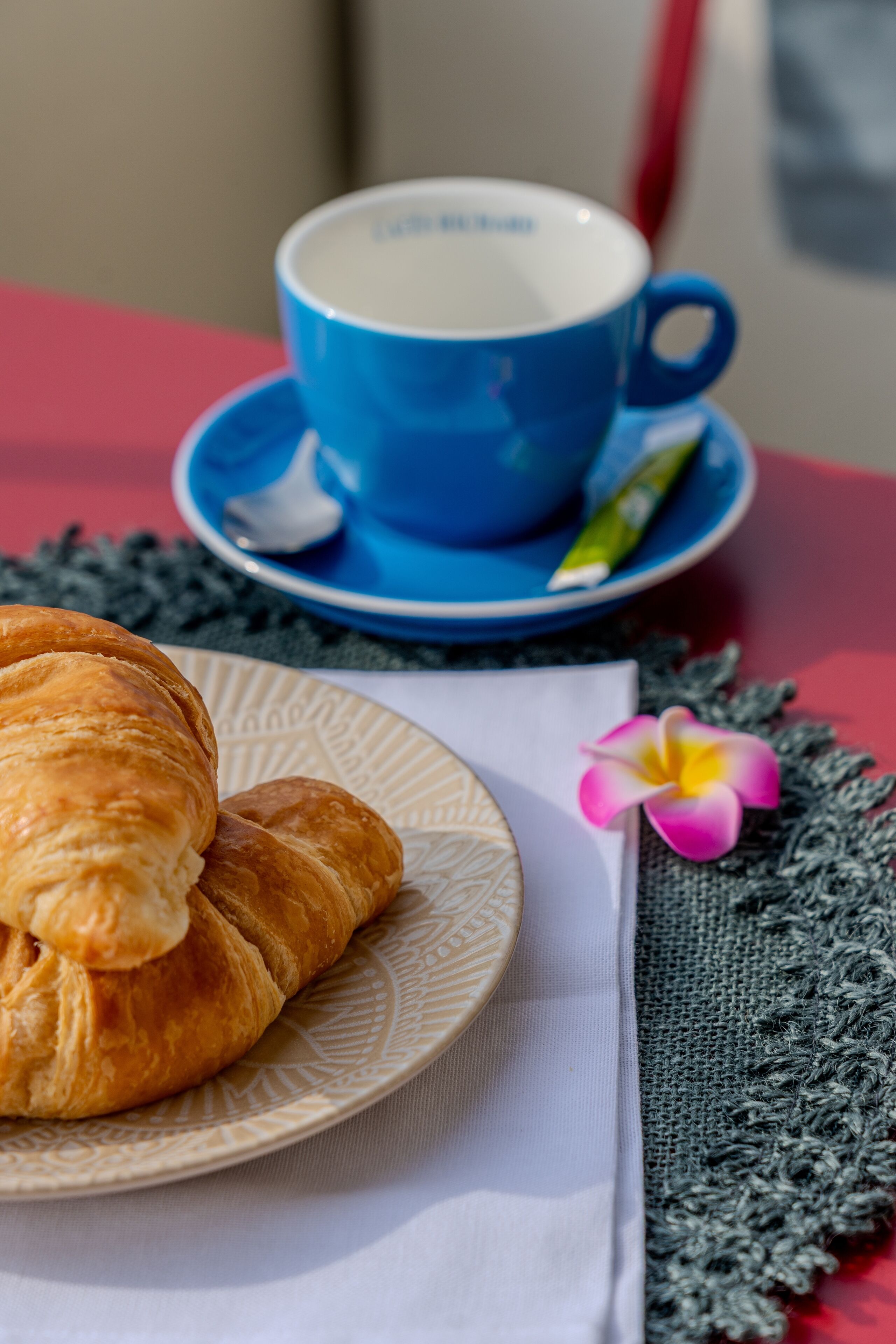 Ein Croissant auf einem Teller, eine blaue Tasse mit Untertasse und eine rosa Blume auf einer Tischdecke.