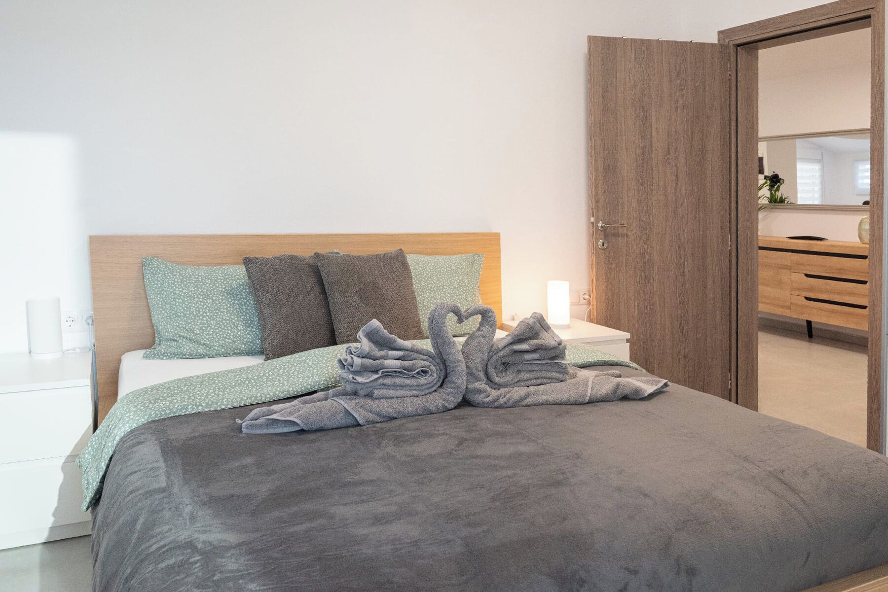 Cama bem arrumada com manta cinza e duas toalhas em formato de cisne, cabeceira de madeira e mesa de cabeceira com abajur.