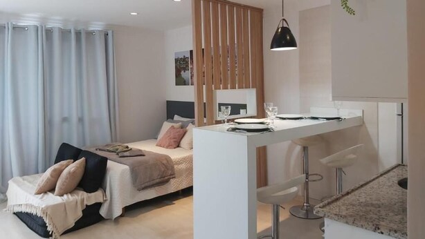 A modern bedroom with a bed, bedside table, and a small sitting area. - Studio Equipado em Apartamento Clube com Academia no Centro de Curitiba MID Home (Centro Curitiba)