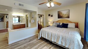 A bedroom with a wooden ceiling fan, a large bed with patterned bedding, a nightstand with a lamp, and a painting on the wall.