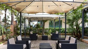 An outdoor seating area with wicker furniture and umbrellas, surrounded by greenery and a paved walkway. - Hotel Villa Merope (Rimini)