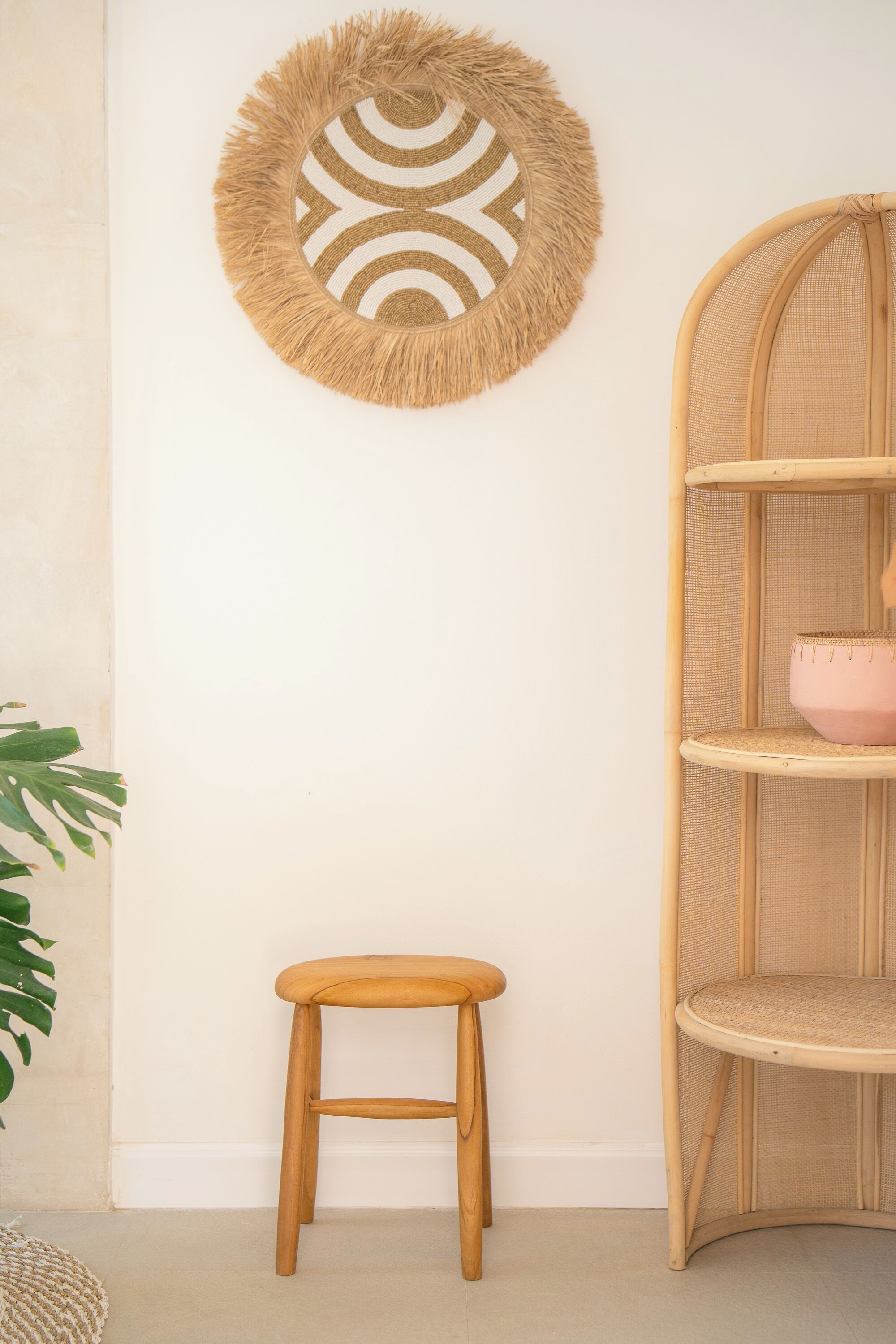 A room with a round woven wall hanging, a wooden stool, and a wicker shelving unit with a pink bowl.