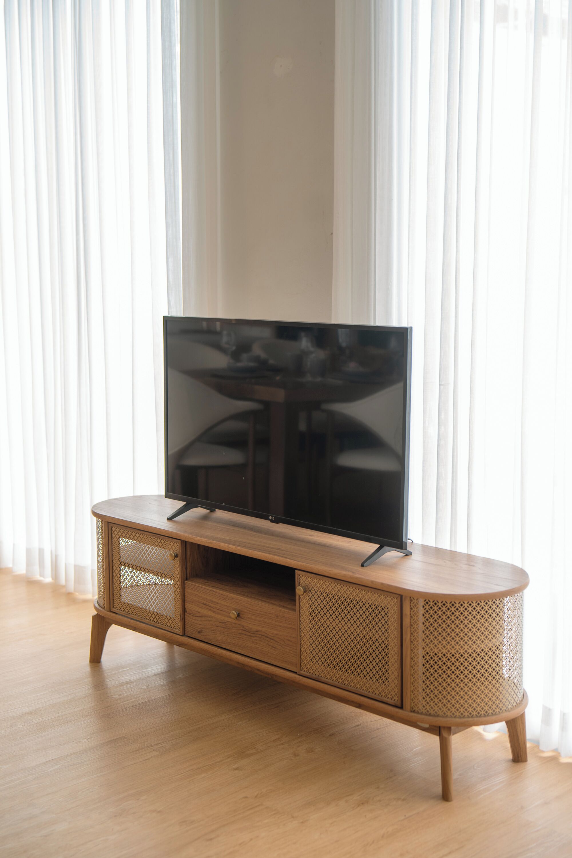 A wooden TV stand with a flat-screen TV, positioned in front of sheer white curtains.