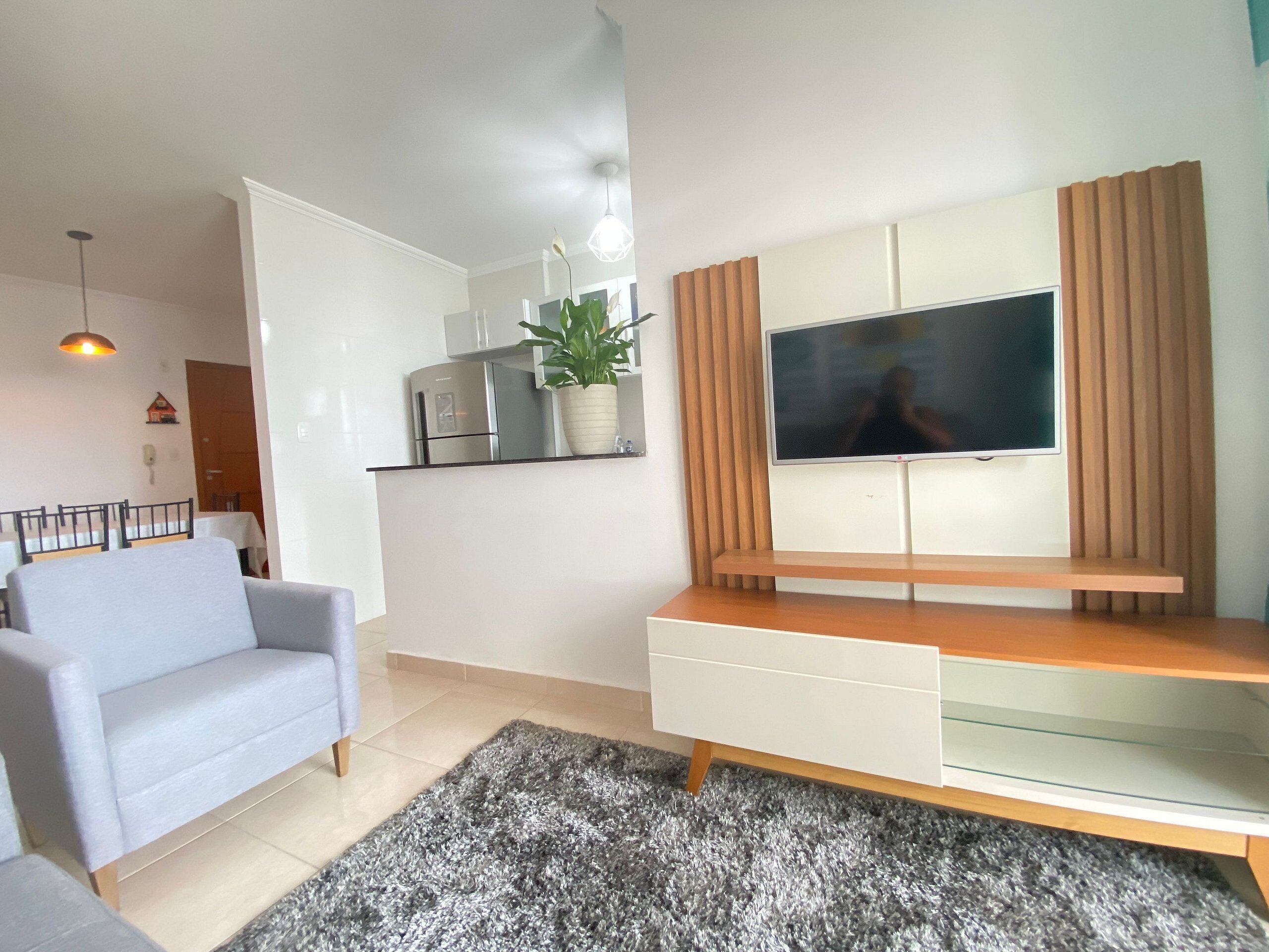 A modern living room with a grey sofa, a TV mounted on a wooden cabinet, a kitchen area with a fridge and a plant, and a tiled floor.