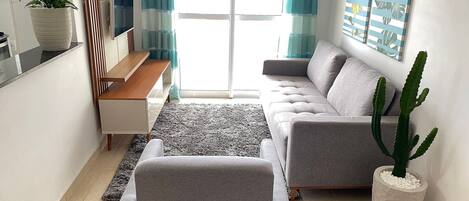 Una sala de estar moderna con un sofá gris, una mesa de centro, una planta en maceta y un ventanal con cortinas.