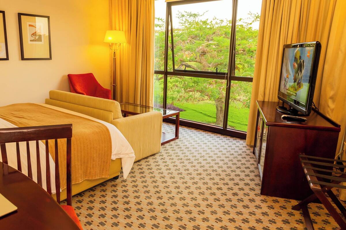 Ein Hotelzimmer mit Bett, Schreibtisch, Stuhl, Fernseher und einem Fenster mit Blick auf Grünflächen.
