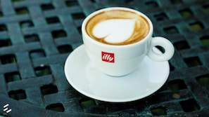 Une tasse blanche ornée d’un motif en forme de cœur réalisé avec de la mousse de lait, posée sur une table en métal verte.