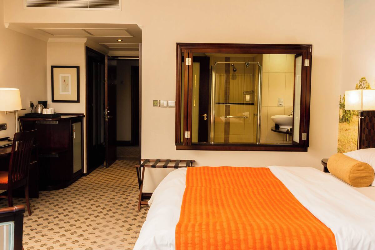 Ein Hotelzimmer mit einem Bett, einem Schreibtisch mit Stuhl, einer Schreibtischlampe, einem Nachttisch und einem Fenster mit Blick auf ein Badezimmer.