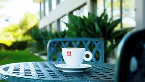 Une tasse blanche ornée d’un logo, posée sur une soucoupe, le tout placé sur une table en métal à l’extérieur.