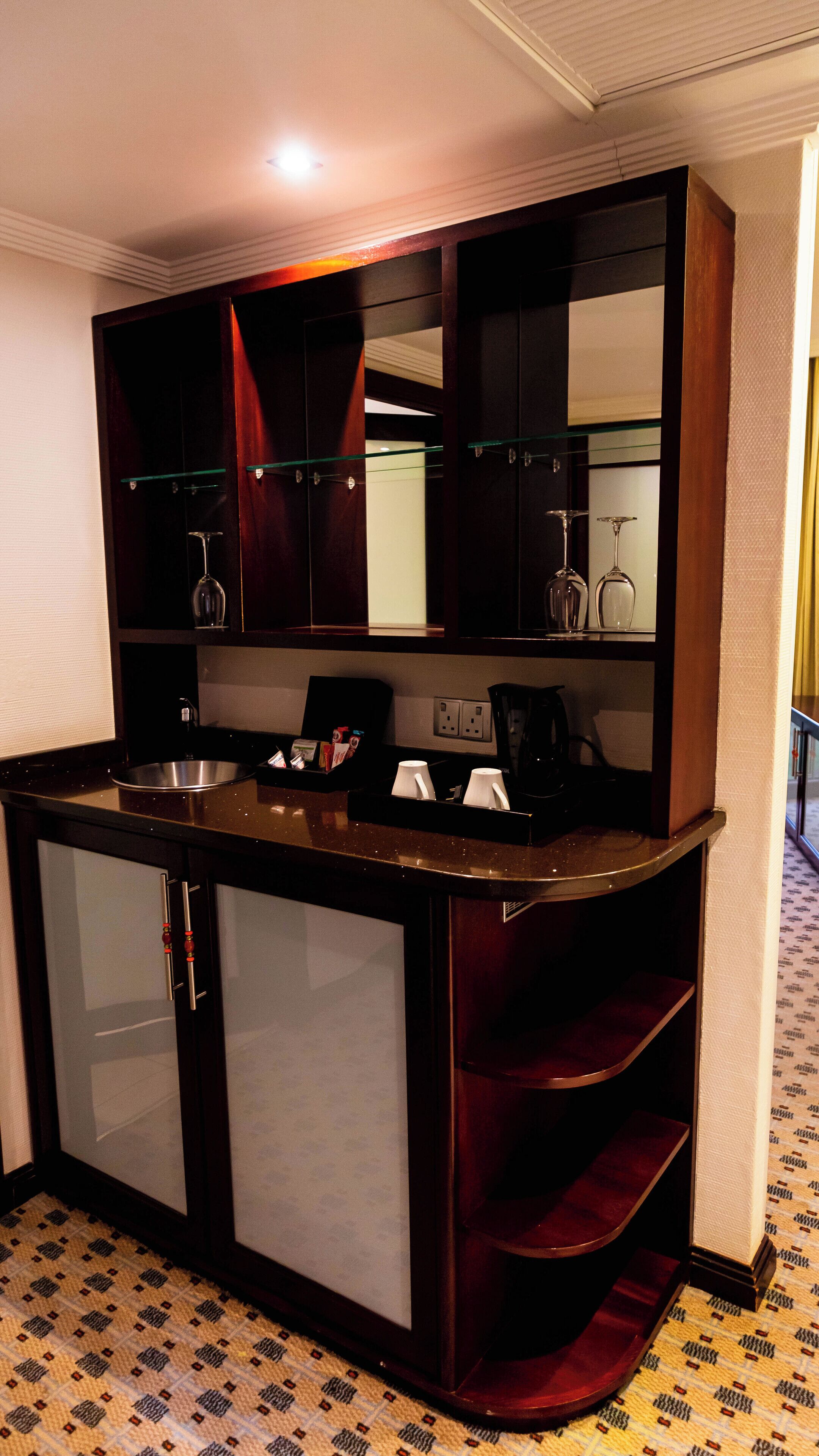 A compact hotel room with a kitchenette, a mirror, and a small seating area with a red cushioned bench.