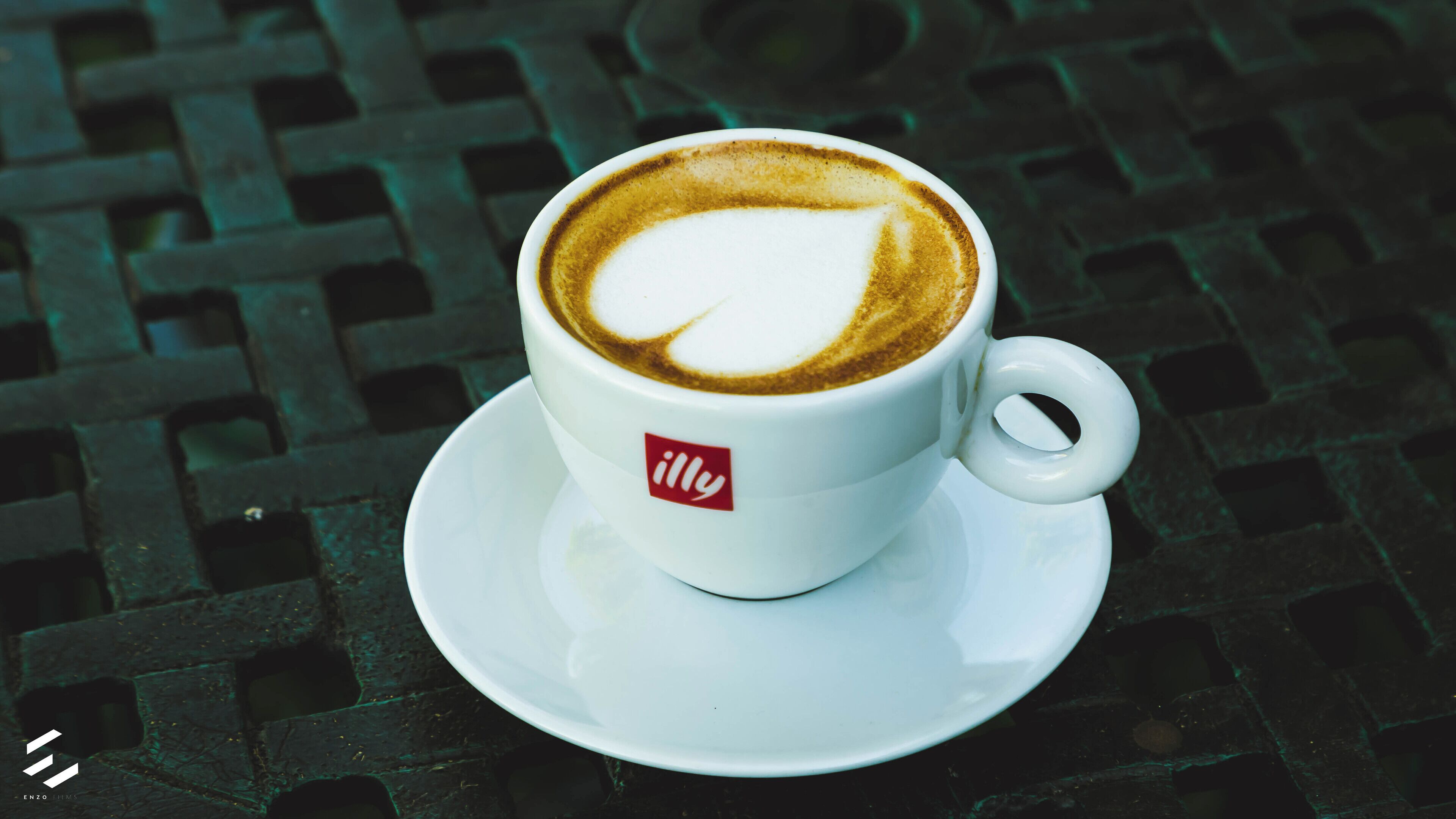 Une tasse de café ornée d’un motif en latte art, posée sur une soucoupe et une tasse blanches, le tout placé sur une table à motifs.