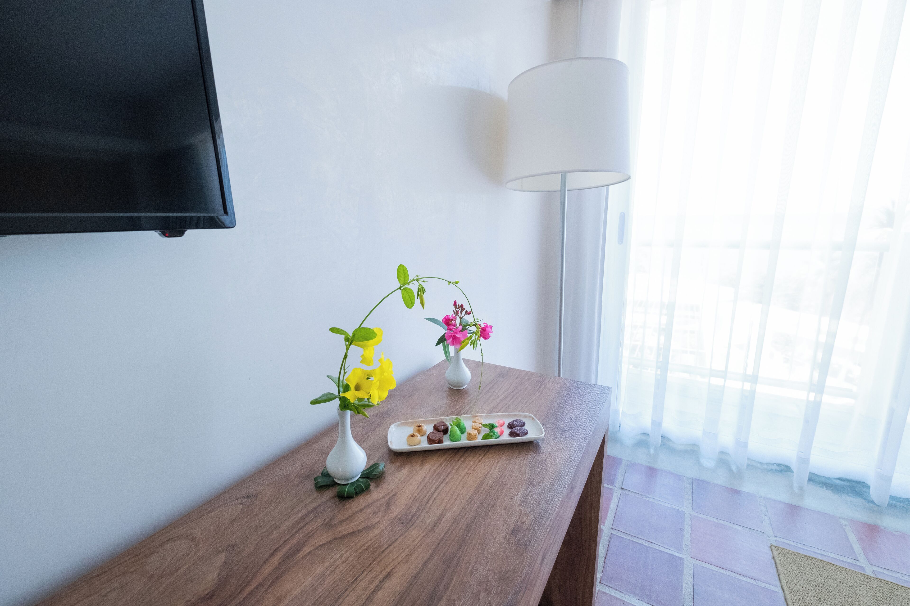 Una mesa de madera con dos jarrones blancos con flores, un plato pequeño con aperitivos, una lámpara blanca y un televisor montado en la pared.