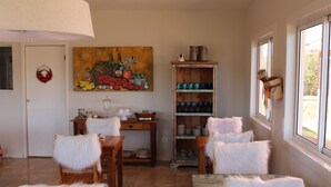 A dining area with a wooden table, chairs with white fur throws, a painting on the wall, and a shelf with dishes and a pitcher. - Vila Chantal by Rivaj  (Campos do Jordão)