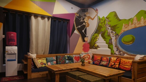 A cozy room with a wooden bench, colorful cushions, a water dispenser, and a mural of a skeleton playing a guitar.