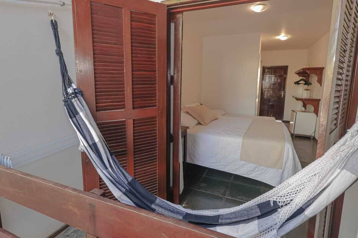 Una hamaca en un vano de puerta, una cama con colcha blanca y color crema, y una puerta de madera con contraventanas.