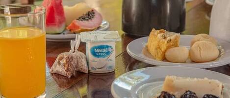 Una mesa de desayuno con un vaso de zumo de naranja, un paquete de Nestlé Grelon, un plato con pan y fruta, y una tetera.