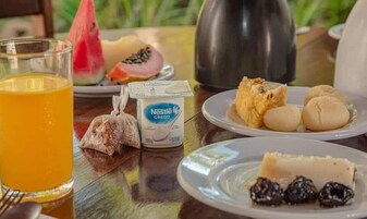 Una mesa de desayuno con un vaso de zumo de naranja, un paquete de Nestlé Grelon, un plato con pan y fruta, y una tetera.