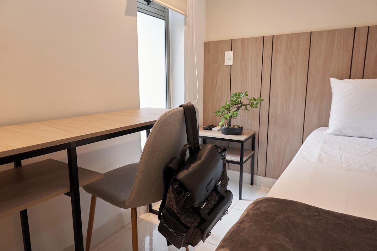 A modern hotel room with a wooden desk, a chair, a small table with a plant, and a bed with a pillow.