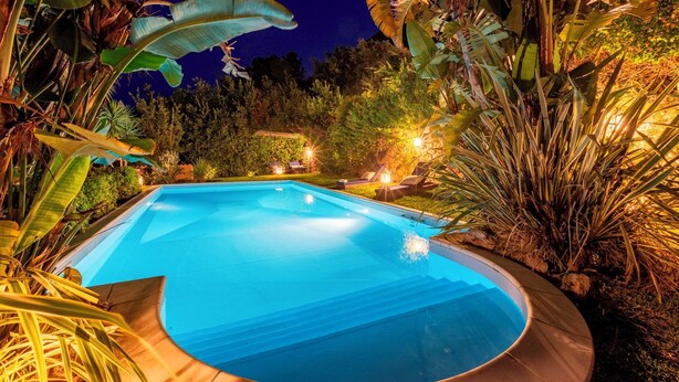 A swimming pool surrounded by tropical plants and trees, with a stone wall and outdoor seating area. - Villa Bambù 9+2, Emma Villas (Maratea)