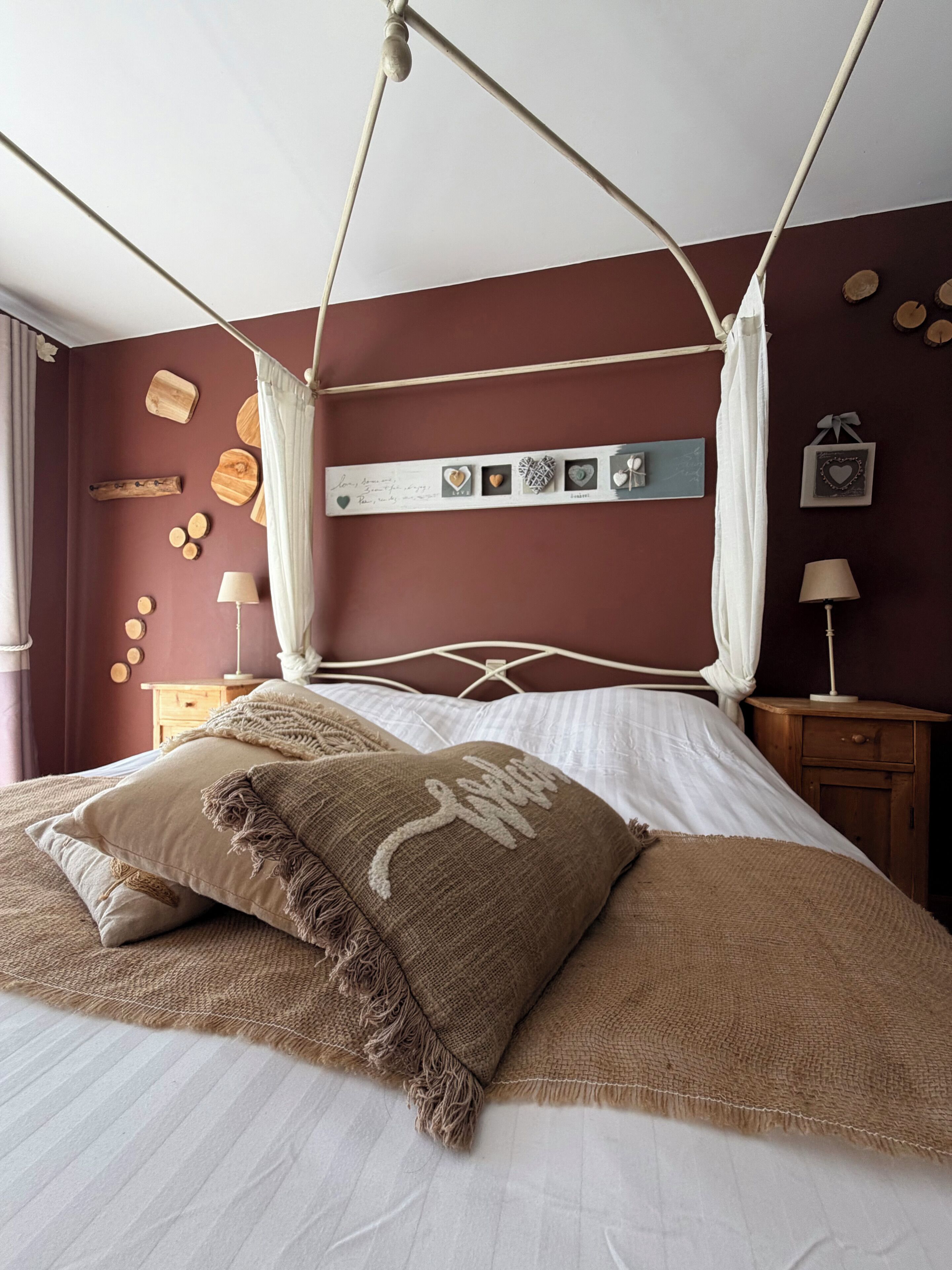 A four-poster canopy bed with a textured throw blanket and pillows, a bedside table with a lamp, and wall decor including wooden plaques and heart-shaped frames.