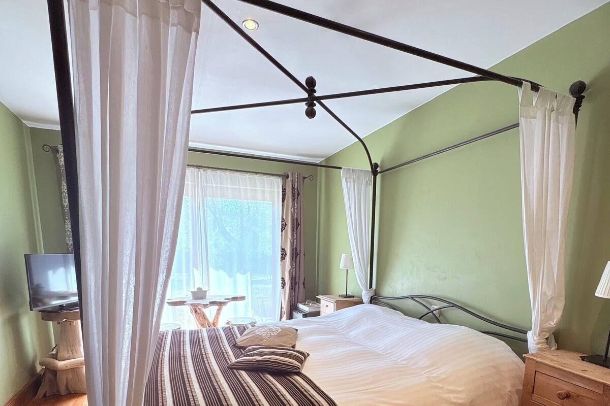 Una cama con dosel con ropa de cama a rayas blancas y marrones, una mesita de noche de madera, una pequeña mesa con una lámpara y un televisor sobre un mueble de madera.
