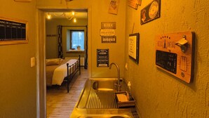 A narrow kitchen with a sink, a stove, and a window. There are signs on the walls and a door leading to another room. - Comme à la ferme (Durbuy)