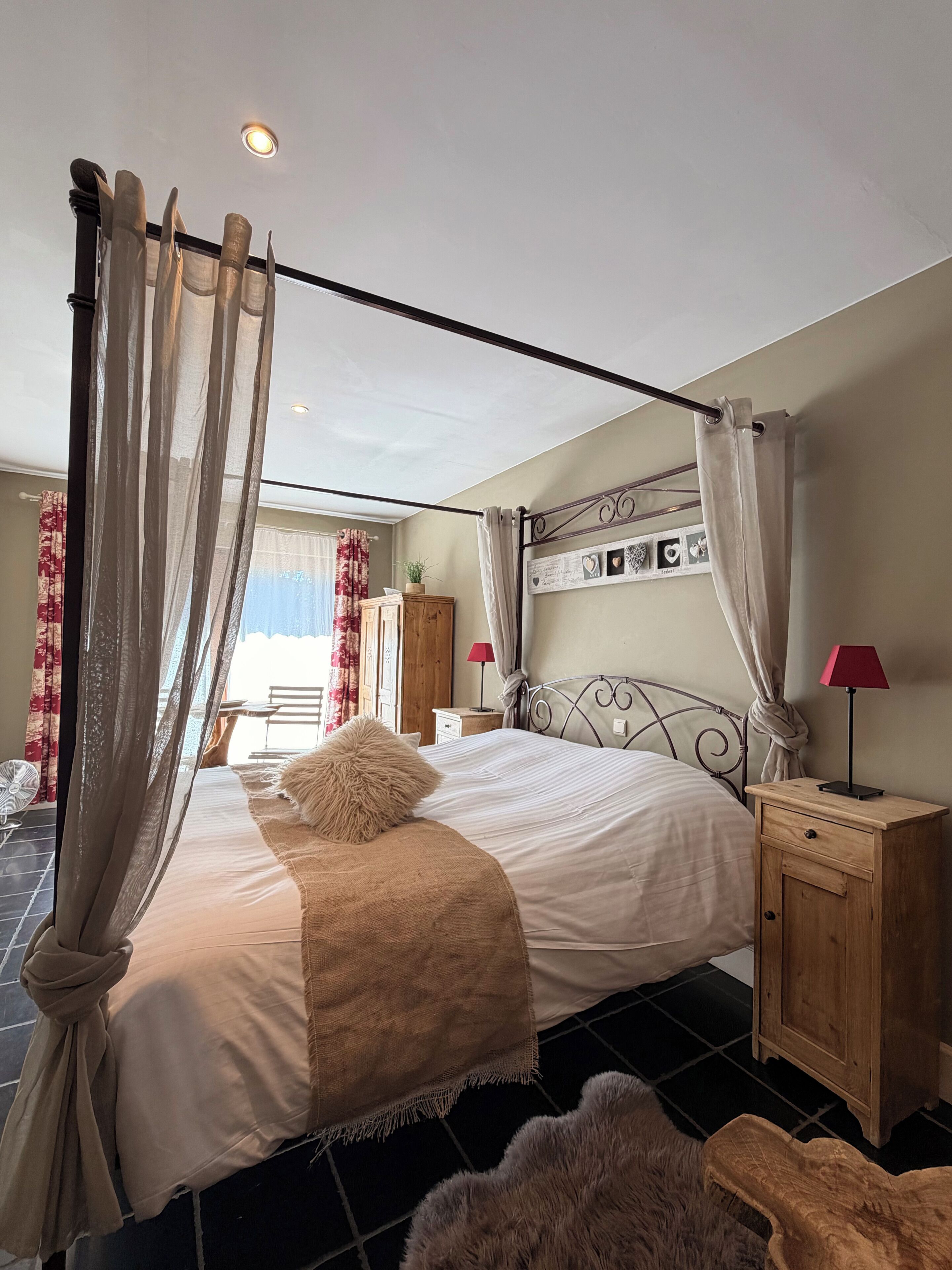 A bedroom with a four-poster bed, a wooden nightstand, a red lamp, and a view of the outdoors.