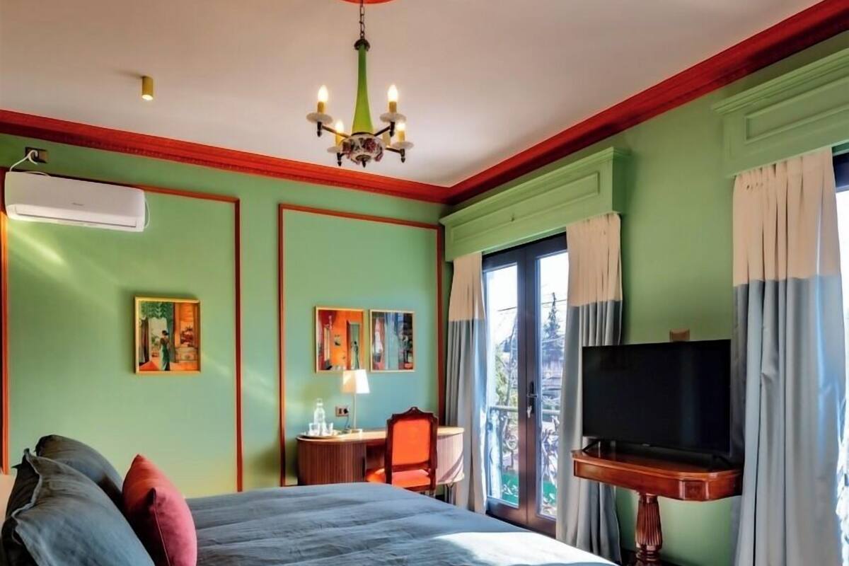Une chambre à coucher avec un grand lit, un téléviseur posé sur un meuble, un bureau avec une chaise et une fenêtre avec des rideaux.