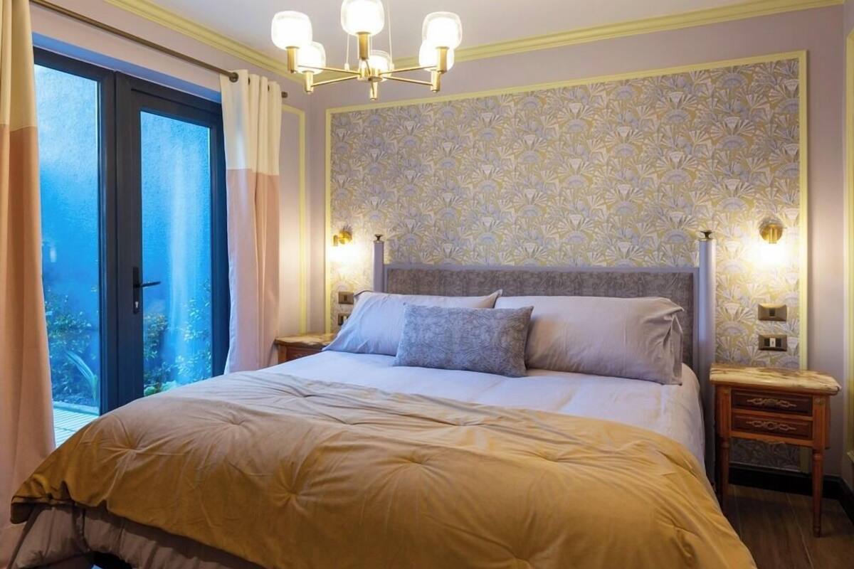 Une chambre à coucher avec un grand lit, un lustre, un papier peint à motifs et une vue sur l’extérieur, visible à travers une porte vitrée.