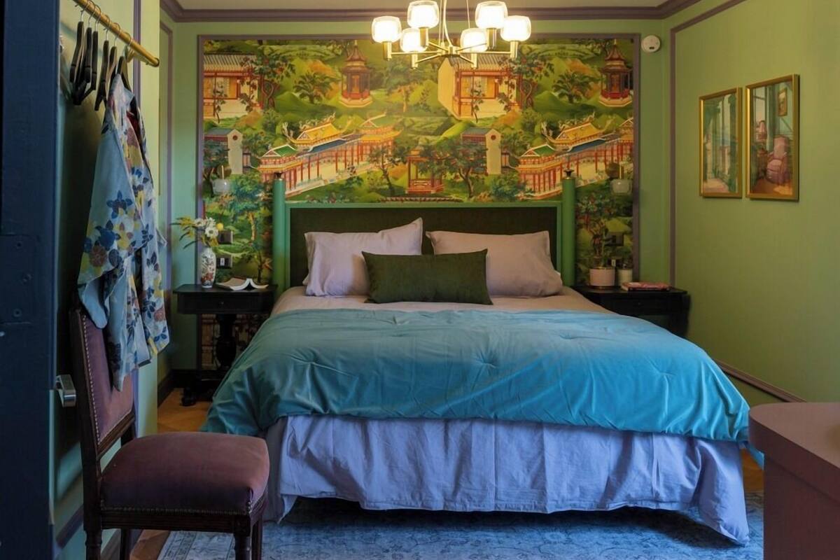 Une chambre à coucher comprenant un lit, une table de chevet, une chaise, un luminaire suspendu et un mur orné d’une fresque.