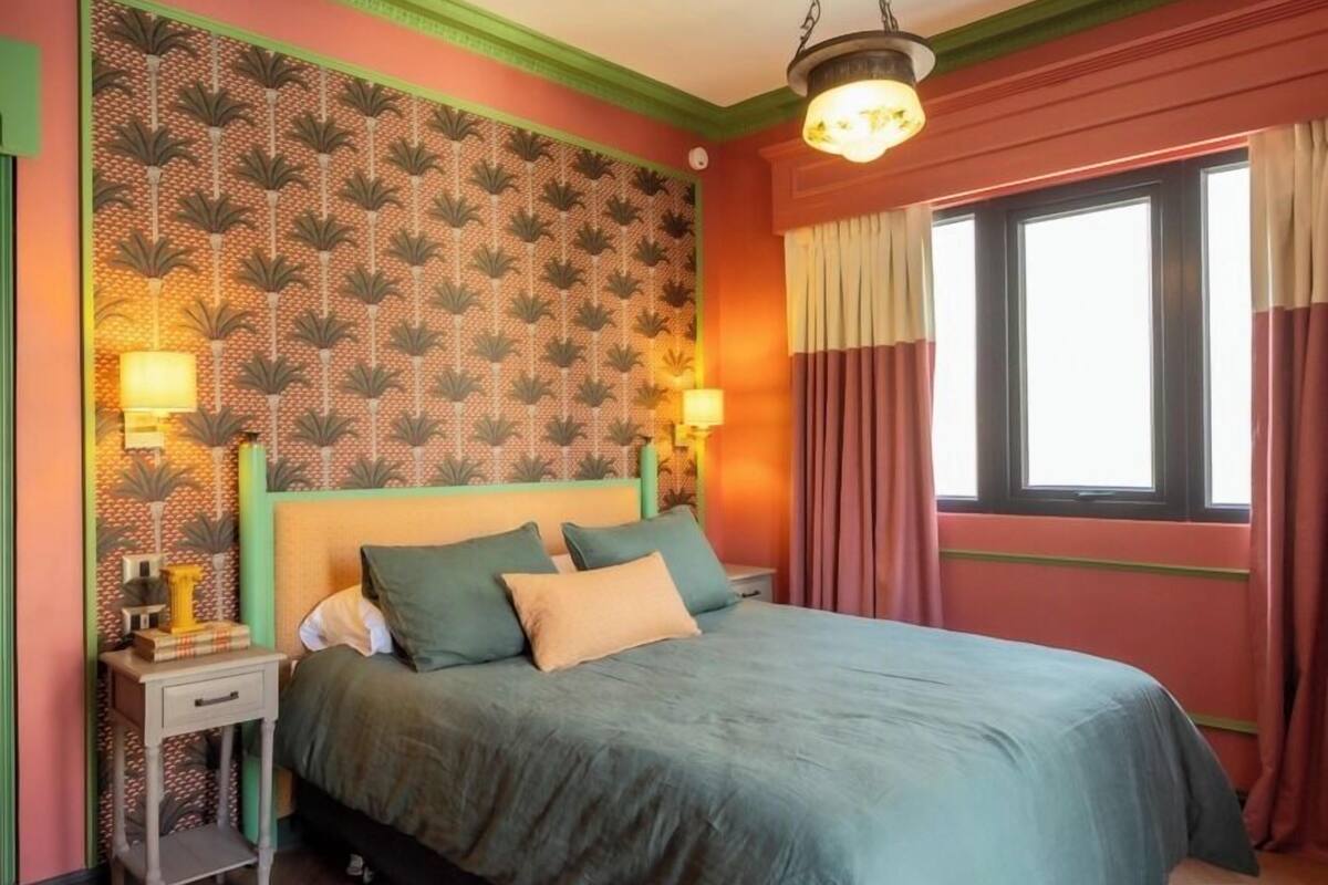 Une chambre à coucher avec un lit, des tables de chevet, un papier peint à motifs, une fenêtre avec des rideaux et un luminaire suspendu.