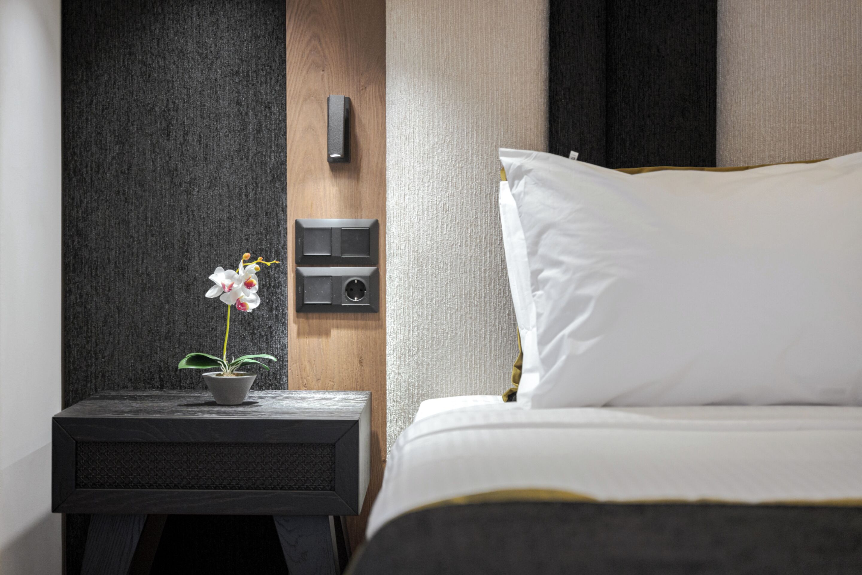 A neatly made bed with white linens and a single white pillow, a small bedside table with a potted orchid, and a wall-mounted charging station.