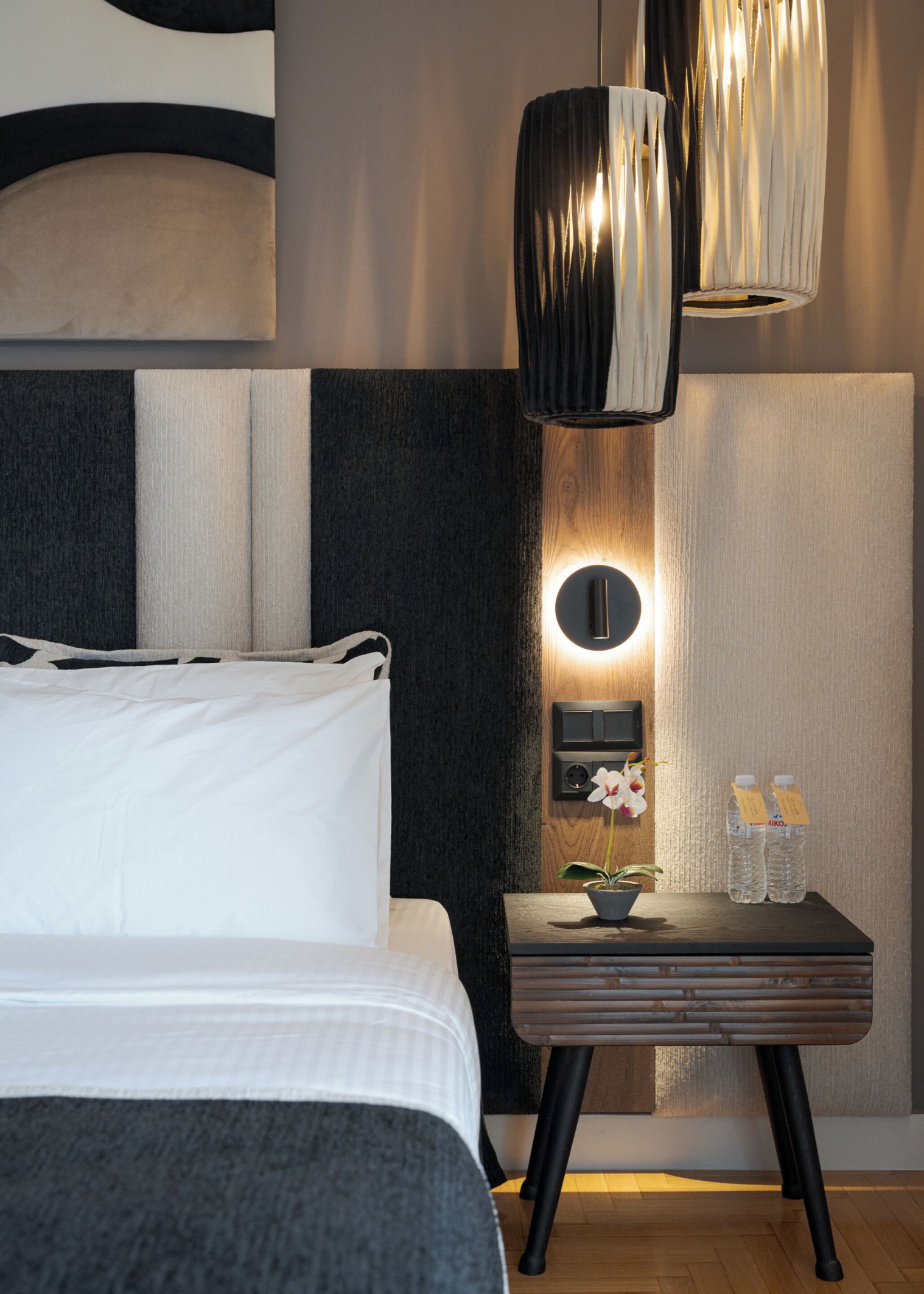 A modern hotel room with a bed, a bedside table with a plant and water bottles, a wall-mounted light fixture, and a wooden panel design.