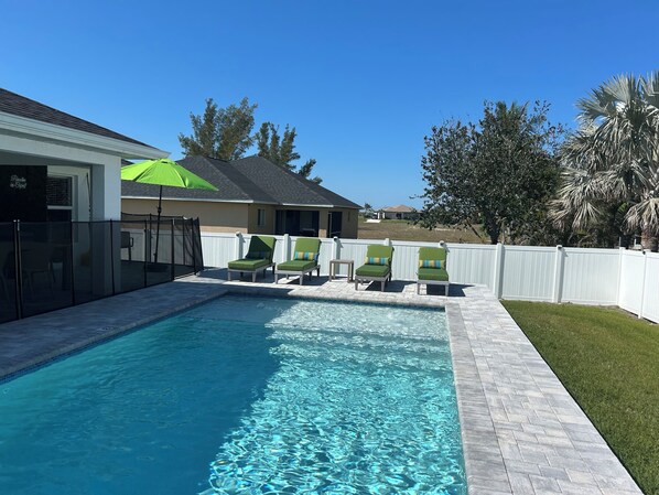 A backyard with a swimming pool, green lounge chairs, a white fence, and a house with a green umbrella.