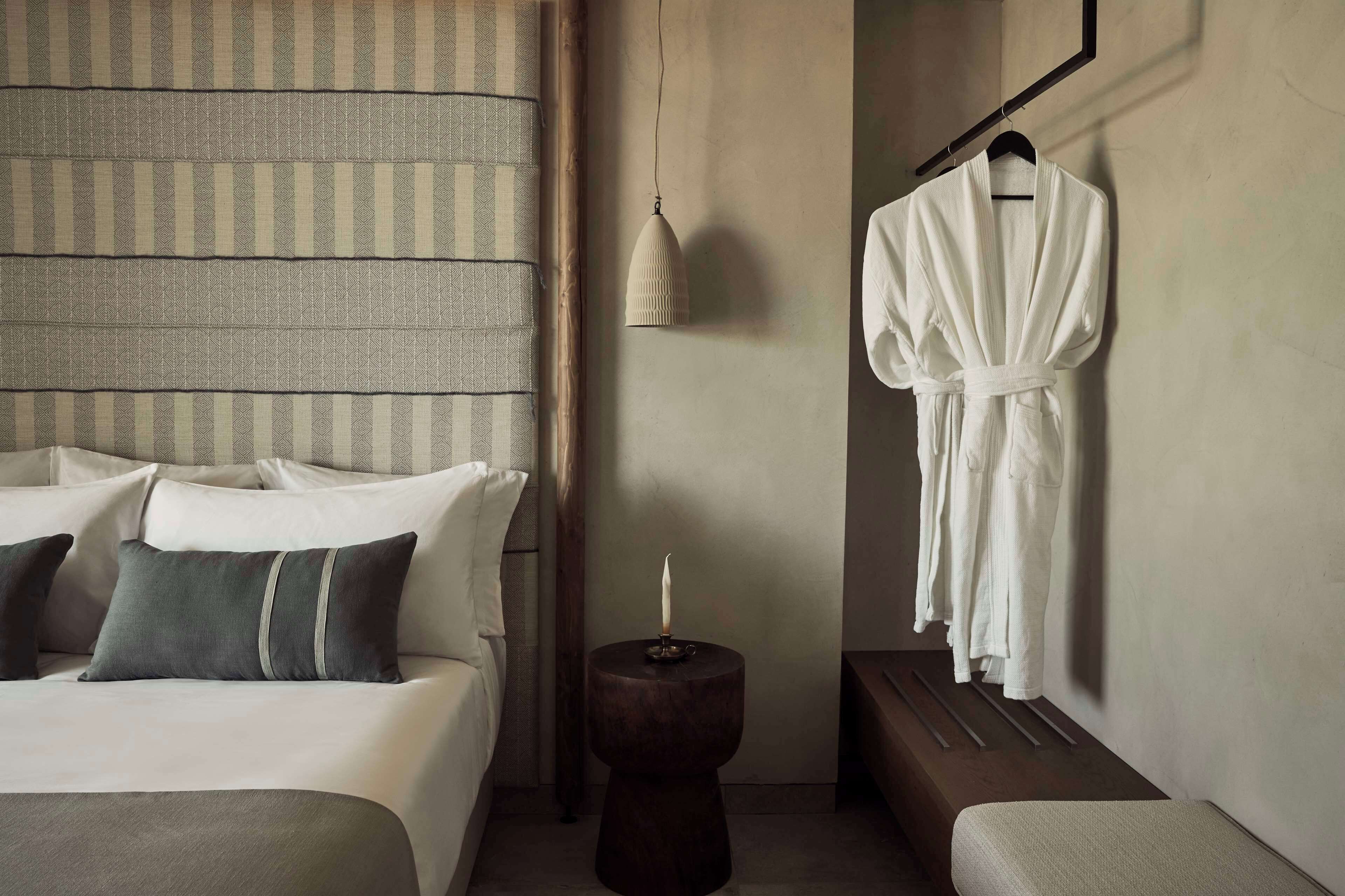 A hotel room with a bed, bedside table, robe hanging, and a wall-mounted light fixture.